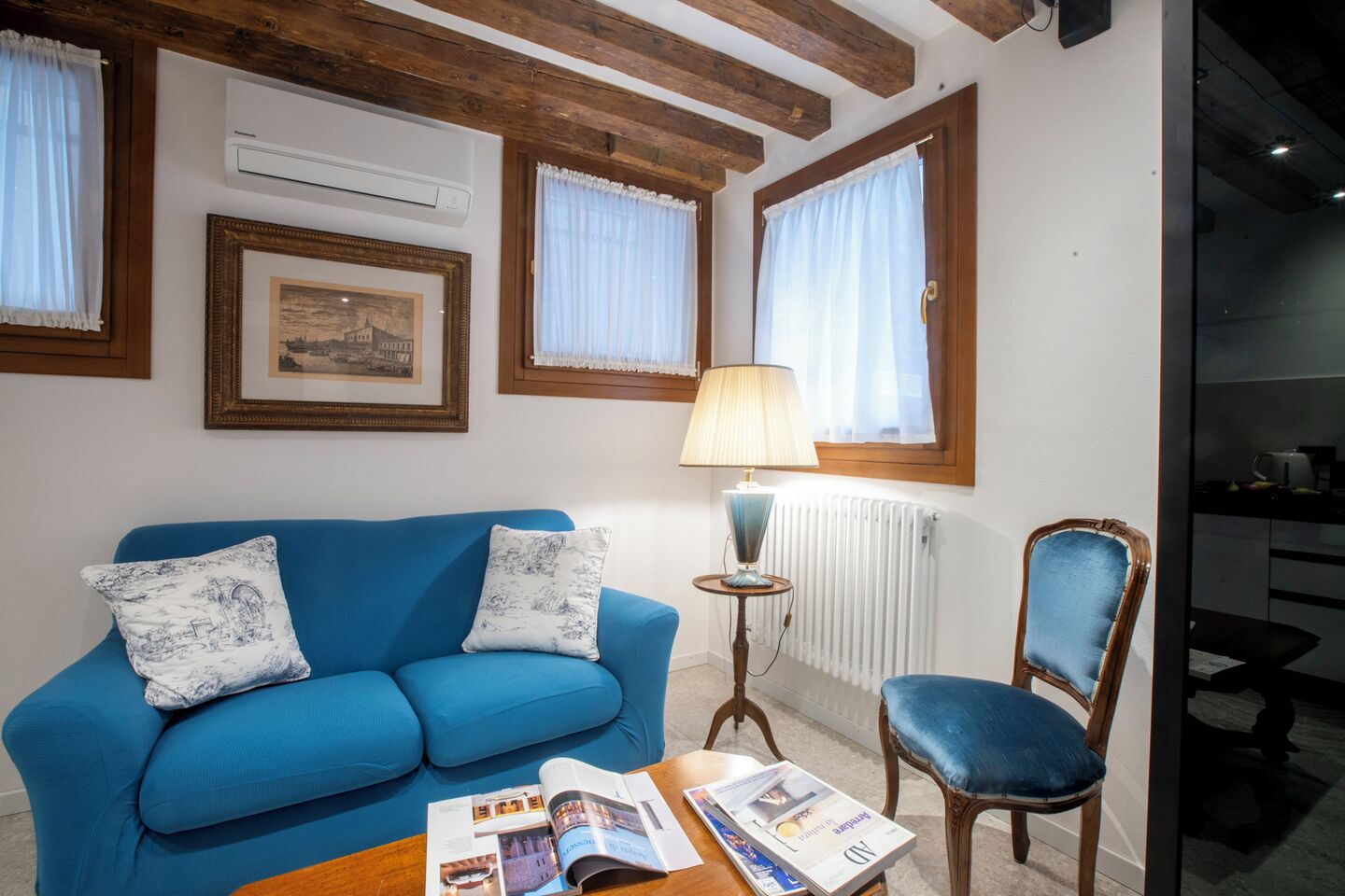 Uma sala de estar com um sofá azul, uma cadeira de madeira, uma mesa com revistas e uma luminária.