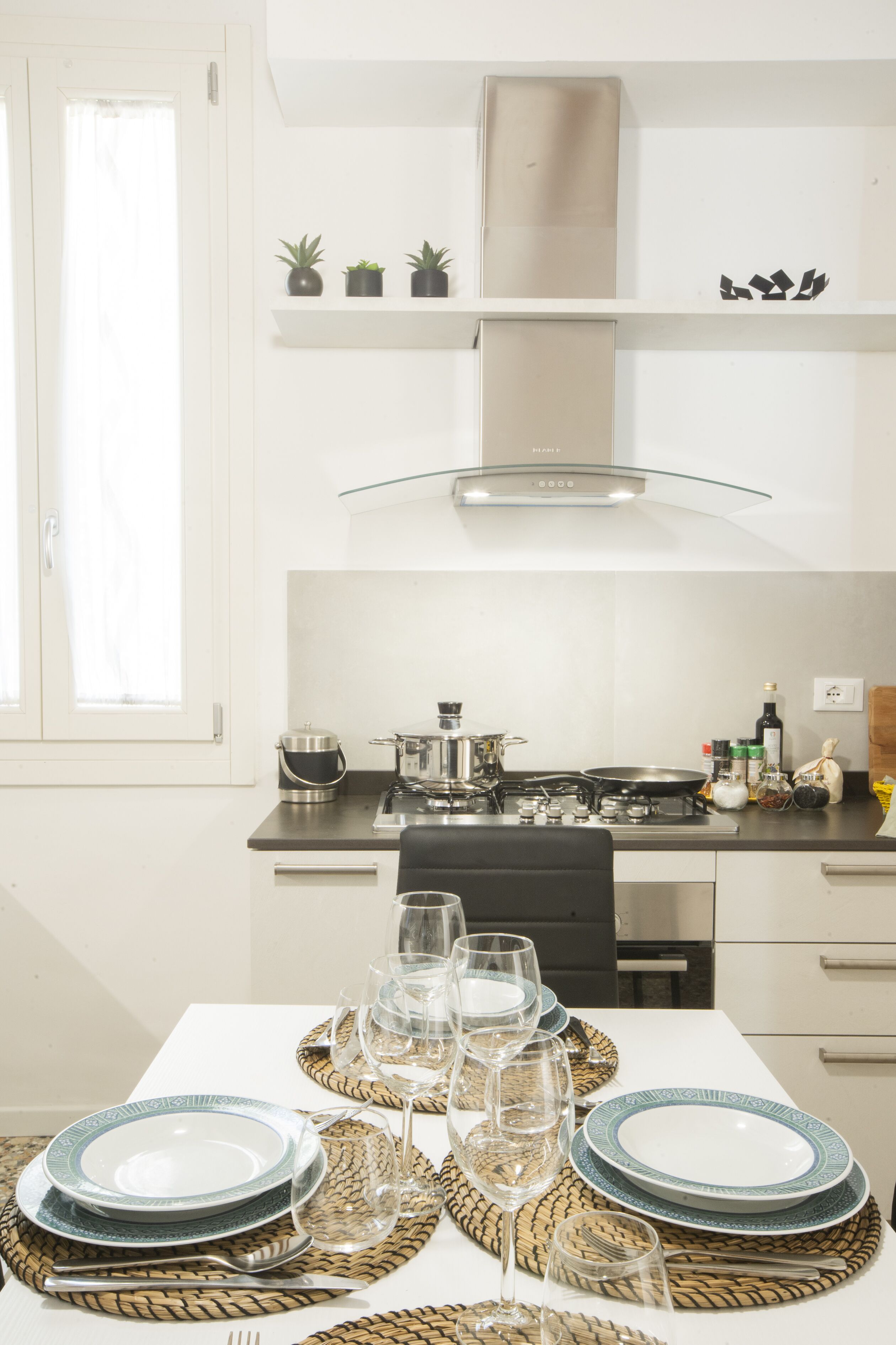 Een moderne keuken met witte kasten, een roestvrijstalen fornuis en een glazen afzuigkap. Boven het fornuis staan drie kleine planten in potten.