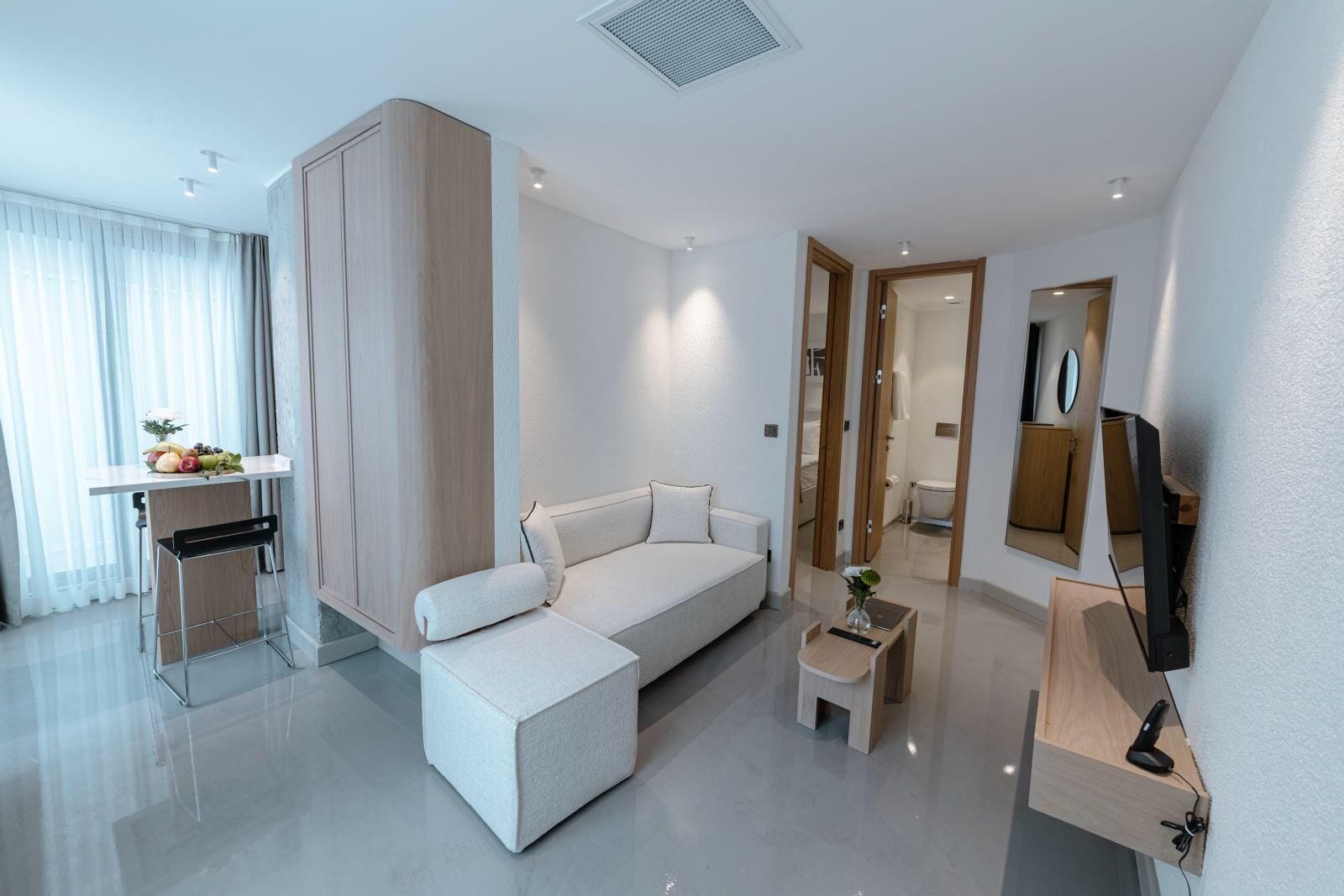 A modern hotel room with a white sofa, a small table with a vase, a TV mounted on the wall, and a mirror.