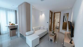 A modern hotel room with a white sofa, a small table with a vase, a TV mounted on the wall, and a mirror.