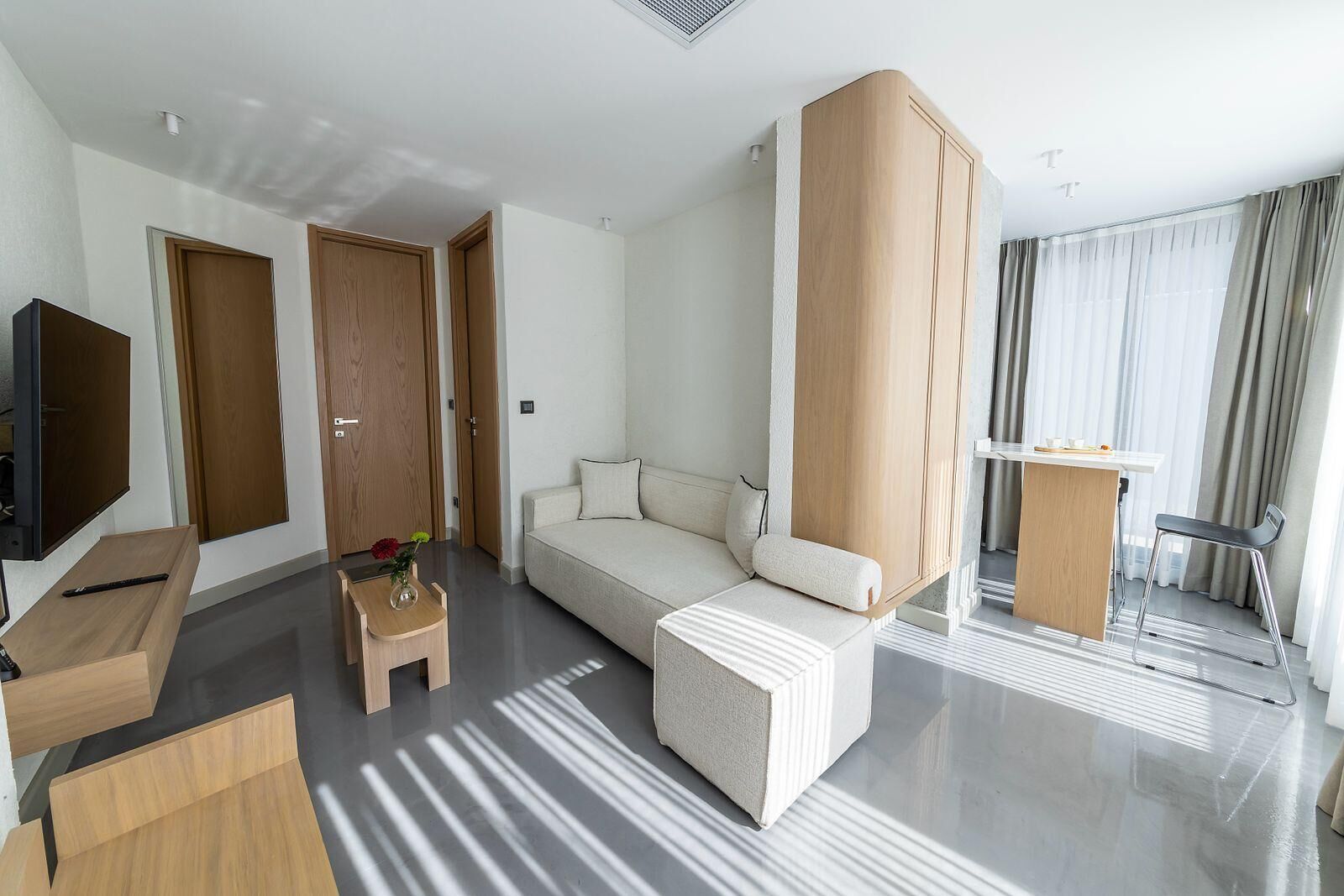 A modern hotel room with a sofa, a small table with a vase, a TV, and a desk with a chair.