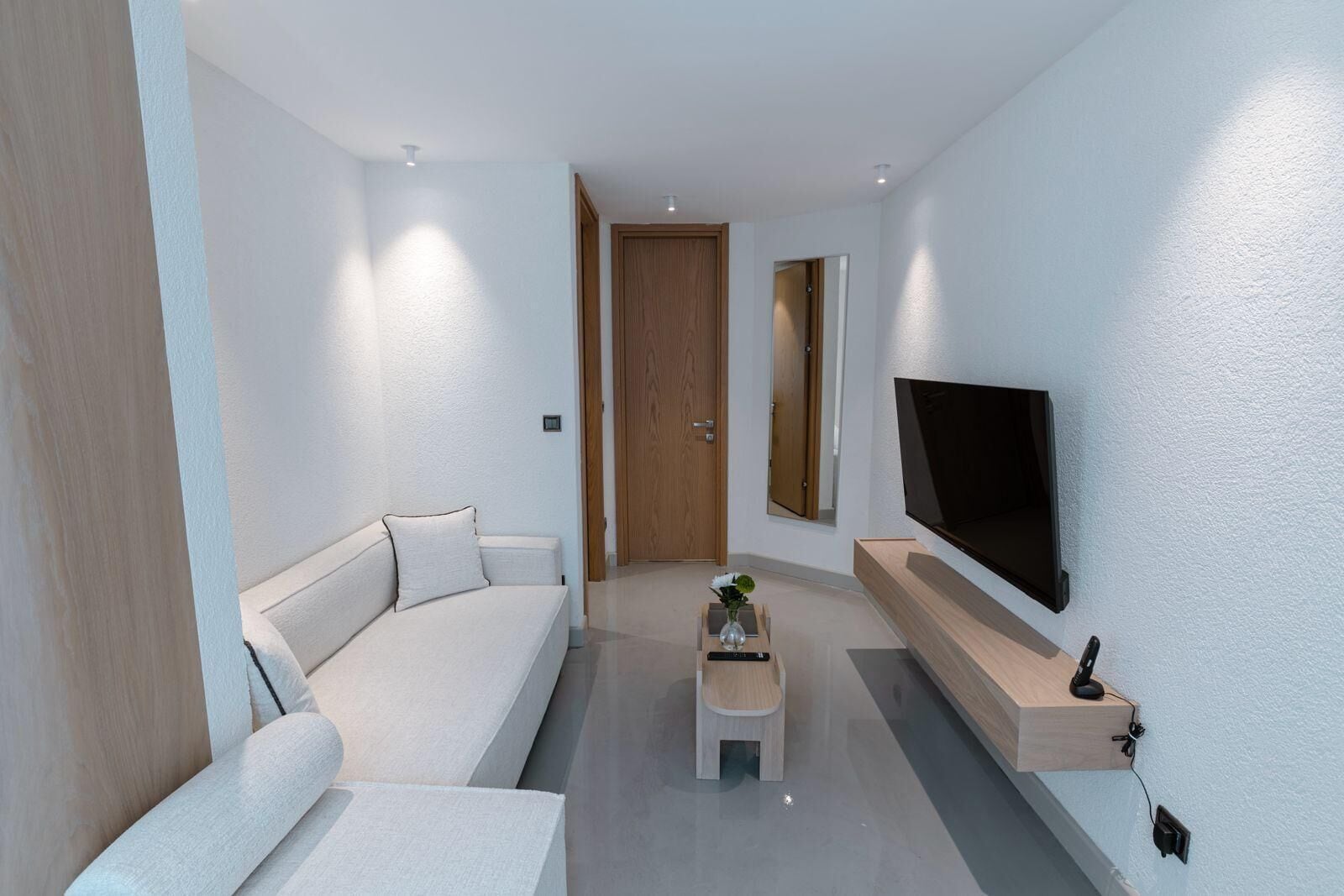 A modern living room with a sofa, a coffee table with a vase, a flat-screen TV mounted on the wall, and a wooden door.