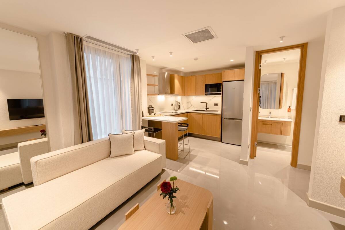 Quarto de hotel moderno com sofá, uma pequena mesa com um vaso, área de cozinha com geladeira e banheiro visível por uma porta aberta.