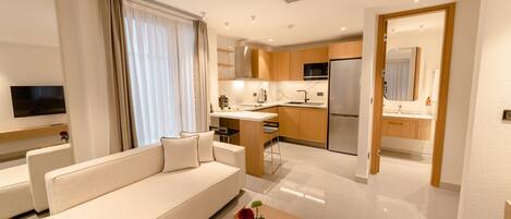 A modern hotel room with a sofa, a small table with a vase, a kitchen area with a refrigerator, and a bathroom visible through an open door.