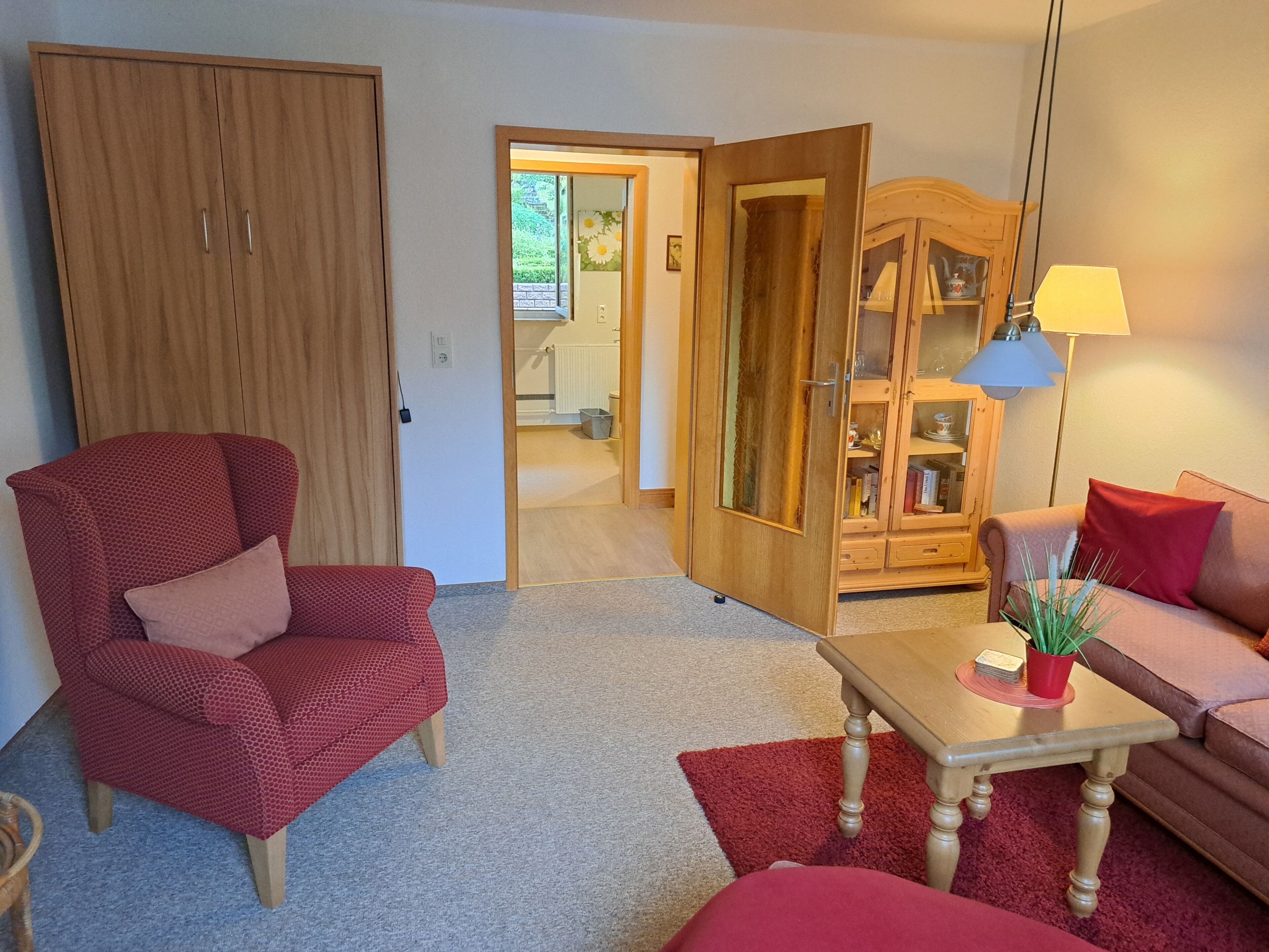 A living room with a red armchair, a wooden cabinet, a table with a lamp, and a red sofa.