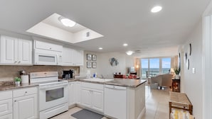 A modern kitchen with white cabinets, a microwave, stove, and a dishwasher. There is a large window providing a view of the outdoors. - Book Now for Fall! Oceanfront! (Panama City Beach)