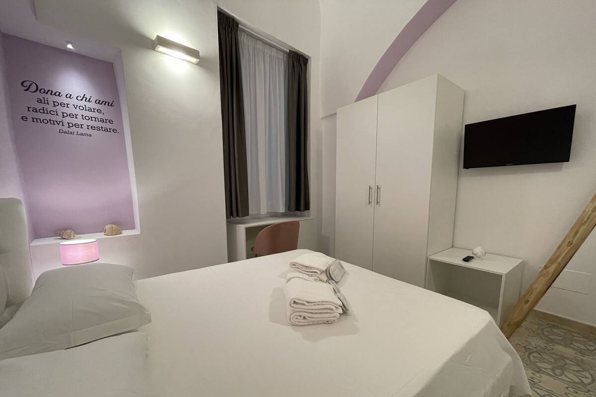 A modern hotel room with a neatly made bed, a wardrobe, a bedside table with a lamp, and a flat-screen TV mounted on the wall.