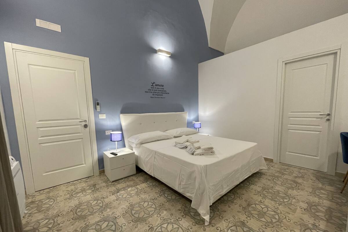 A bedroom with a bed, two bedside tables, a wall-mounted light, and a door.