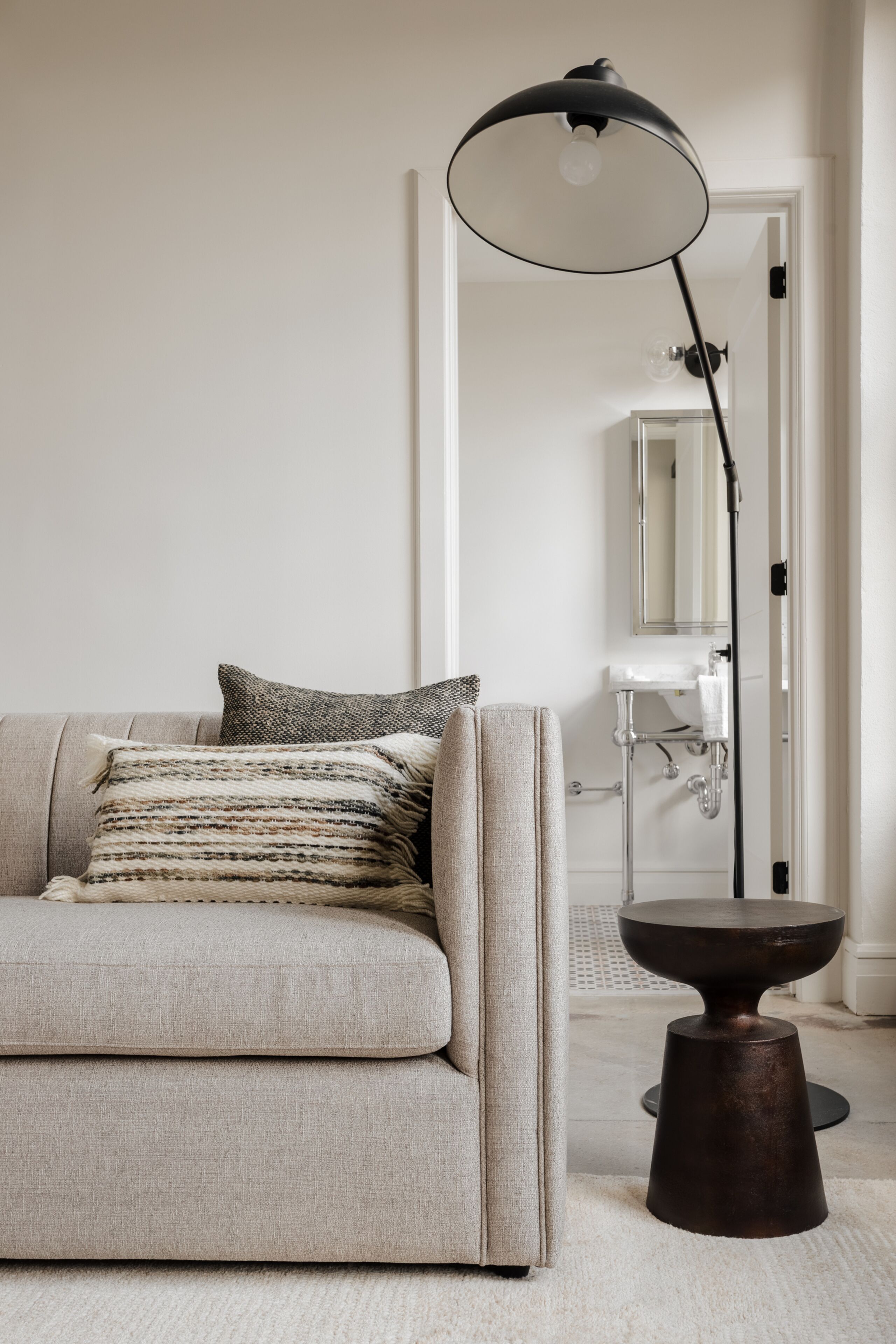 A neutral-toned room with a sofa, a floor lamp, a small table, and a bathroom visible through an open door.