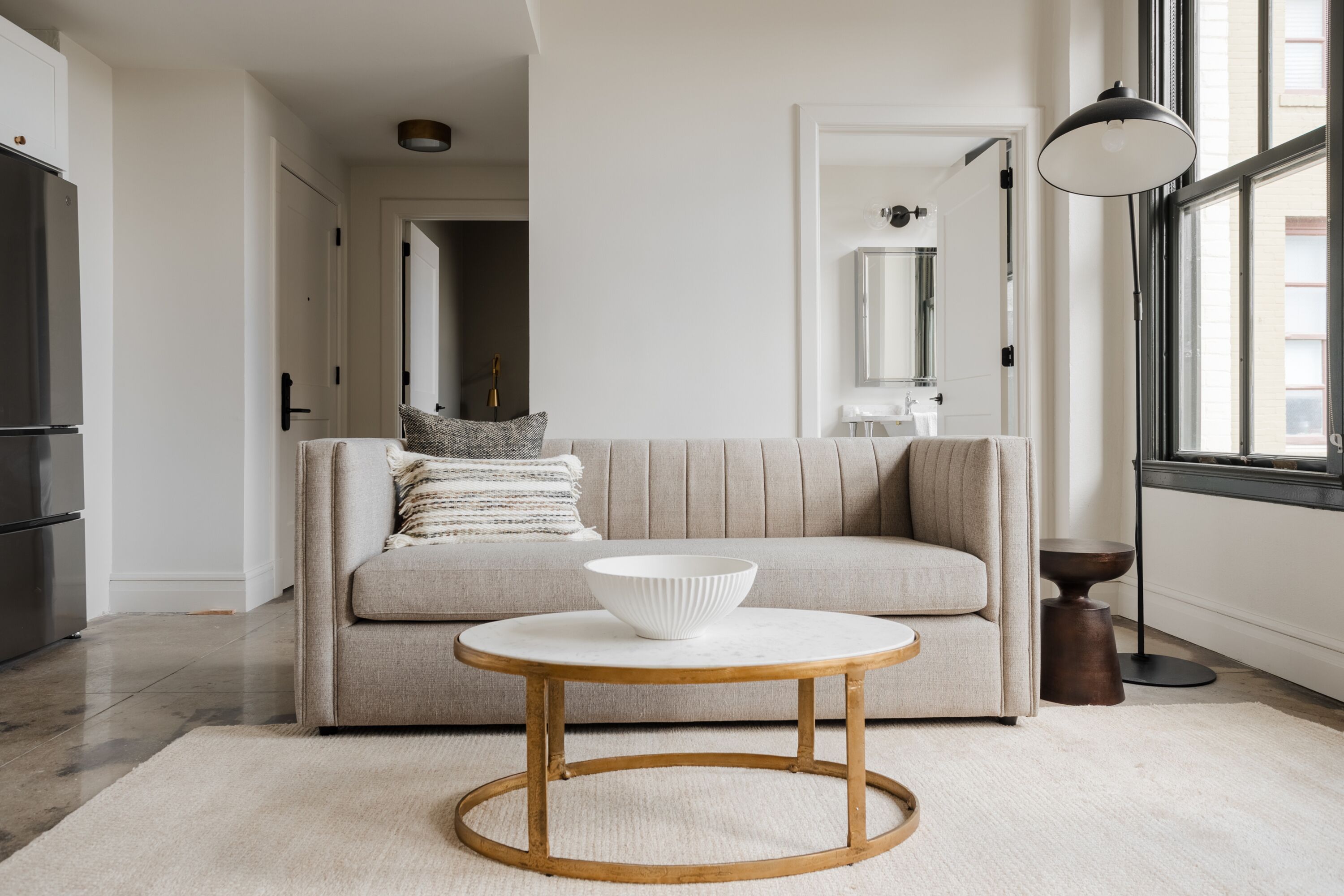 A modern living room with a beige sofa, a round wooden coffee table, and a large window providing natural light.