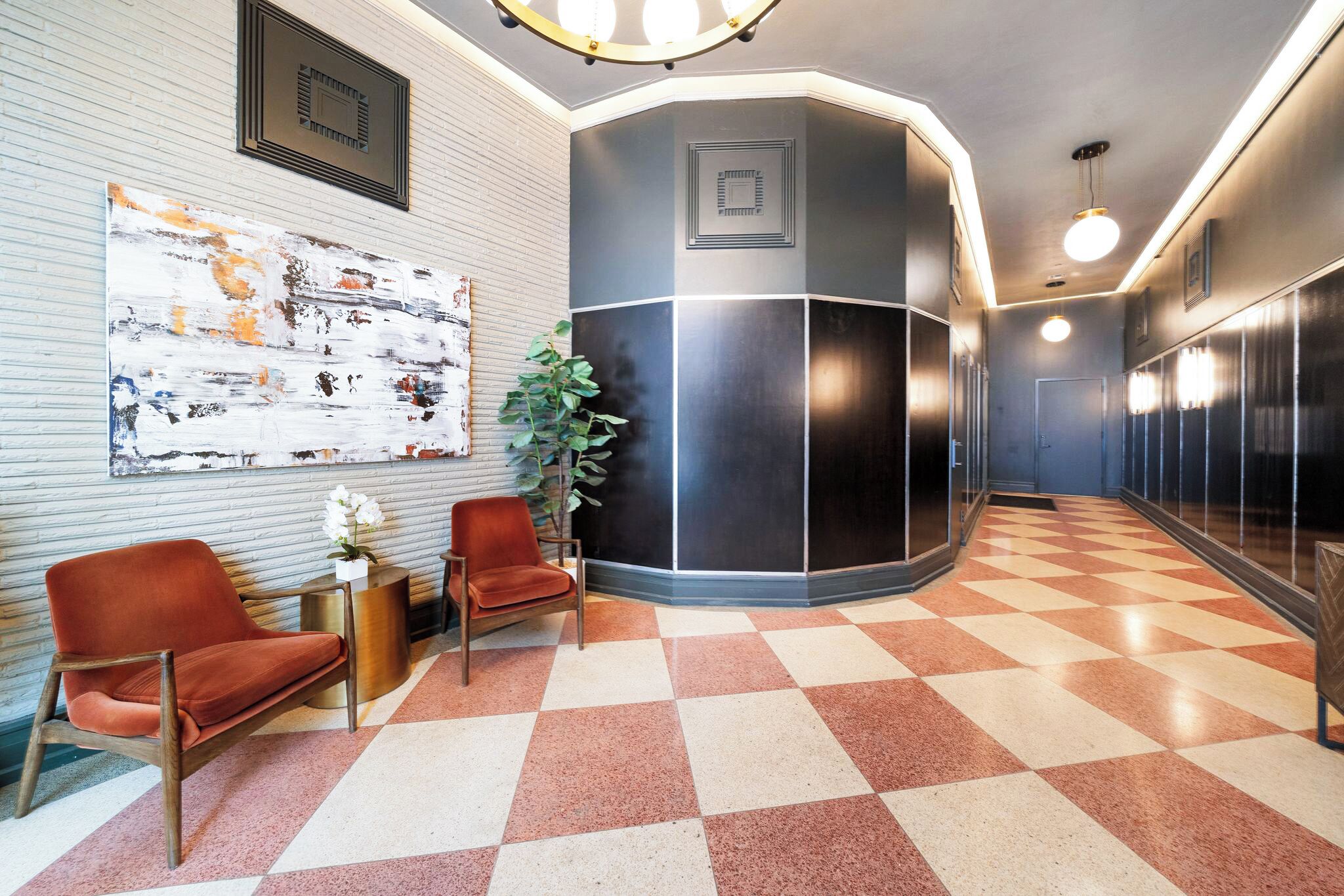 A lobby with a checkered floor, two red chairs, a large abstract painting, and a potted plant.