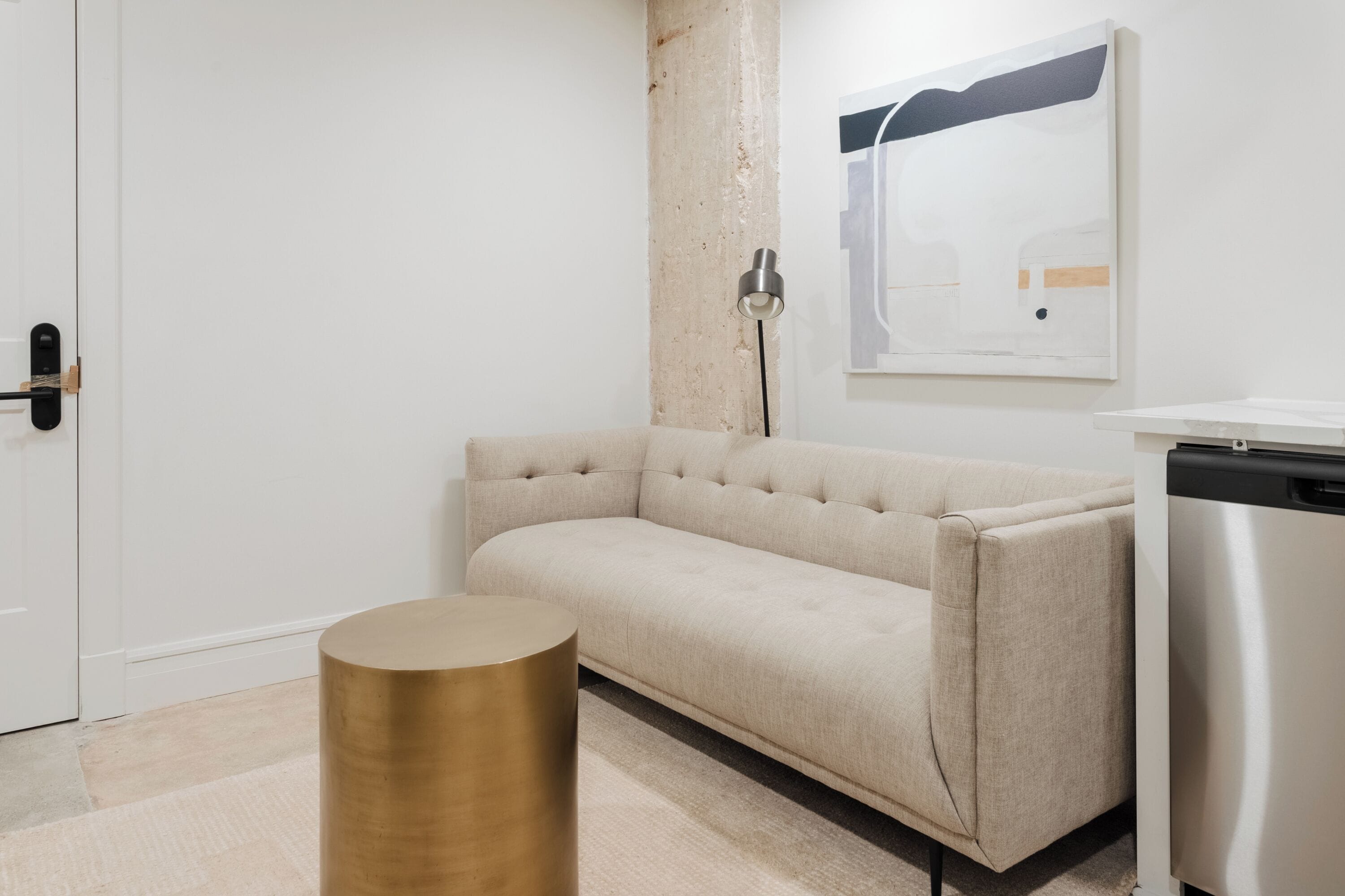 A modern, minimalist room with a beige sofa, a round gold side table, a white door, and a stainless steel appliance.