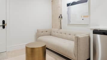 A modern, minimalist room with a beige sofa, a round gold side table, a white door, and a stainless steel appliance.