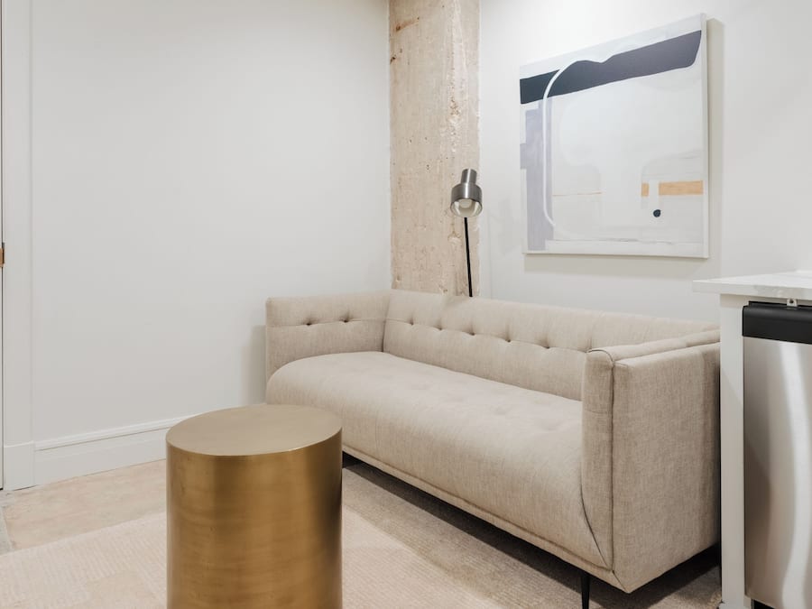 Quarto moderno e minimalista com sofá bege, mesa lateral redonda dourada, porta branca e eletrodoméstico de aço inoxidável.