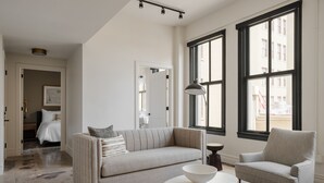 Un saló modern amb un sofà, una butaca i una taula rodona amb un bol. Hi ha finestres grans i una porta que condueix a una altra habitació.
