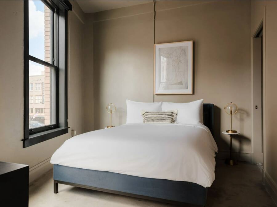 A modern bedroom with a large bed, a window, a framed artwork, and two bedside lamps.