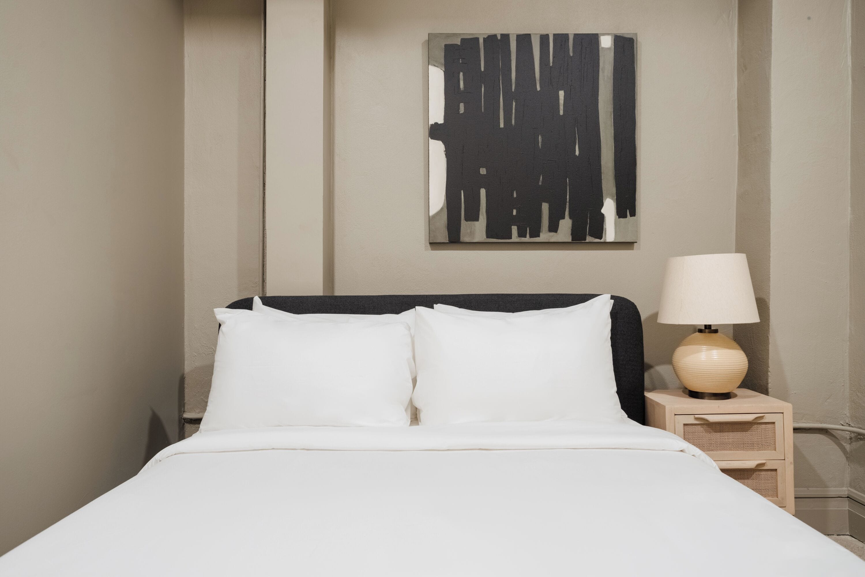 Una cama bien hecha con sábanas blancas, una lámpara de noche y una obra de arte abstracta en la pared.