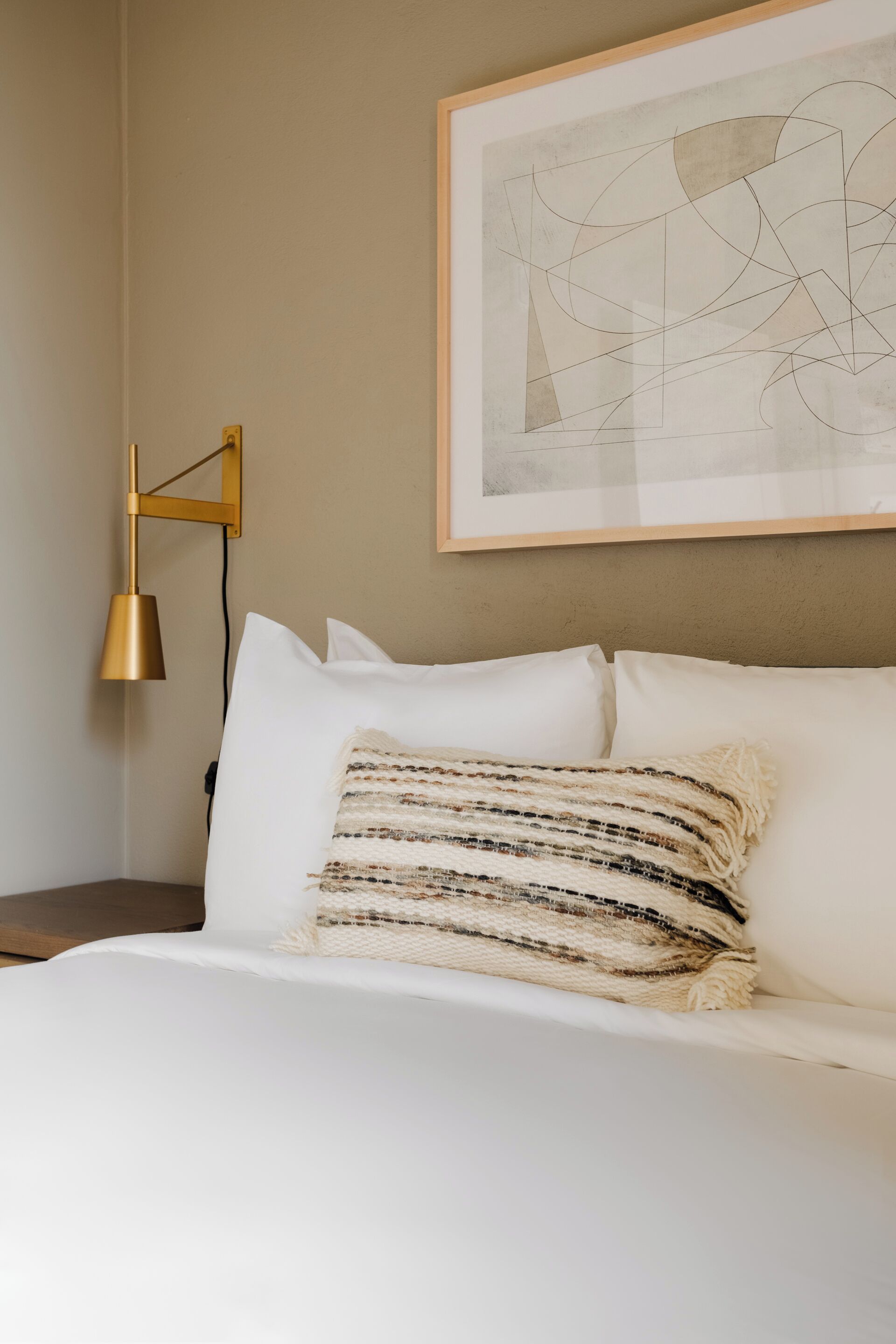 Een net opgemaakt bed met witte lakens en een gestreept kussen, een gouden vloerlamp en een lijst met een abstract schilderij aan de muur.