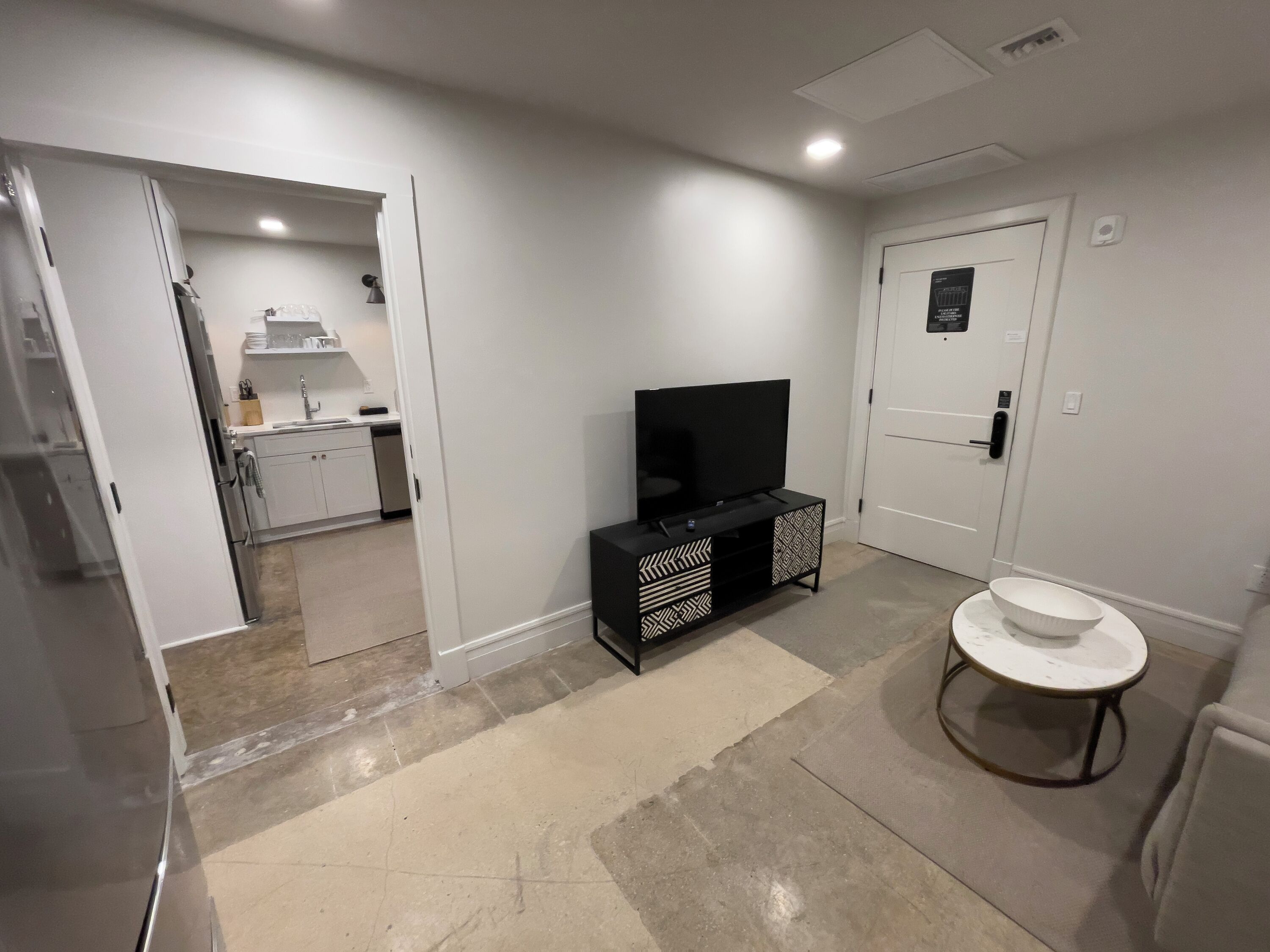 Una habitación moderna y luminosa con una televisión sobre un mueble negro, una mesita redonda con un cuenco blanco, y una cocina visible a través de una puerta abierta.