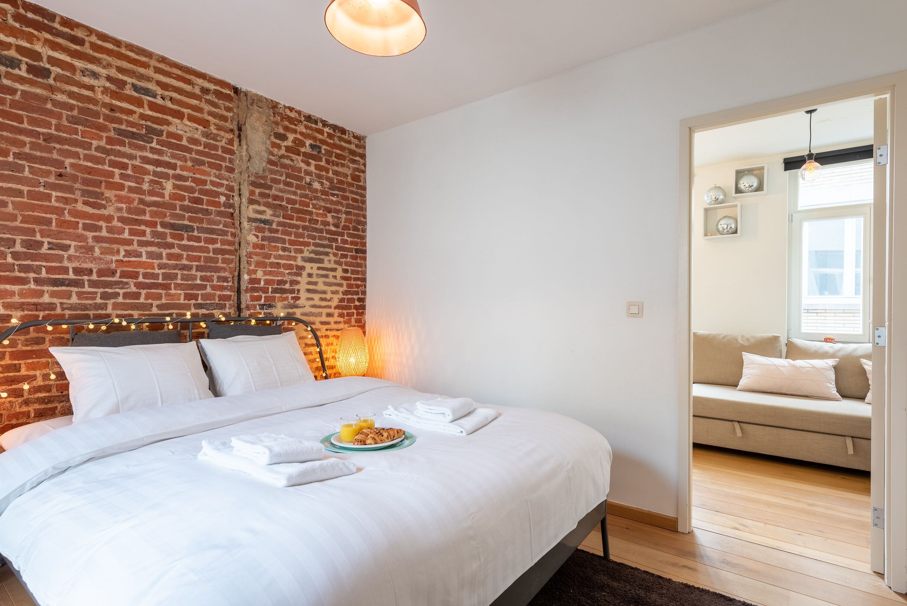 Quarto com parede de tijolos, cama com roupa branca, mesa de cabeceira com abajur e vista para uma sala de estar com sofá.