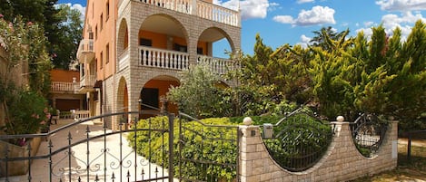 A multi-story building with a balcony, a gated entrance, and surrounding greenery.