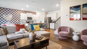A modern living room with a sofa, coffee table, and chairs, a kitchen with white cabinets and appliances, and a staircase leading to another level. - 6min to UNCC: Spacious 5bd with Games Backyard (Charlotte)