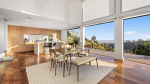 A modern kitchen with wooden cabinets, a dining area with a table and chairs, and large windows offering a view of a hilly landscape. - Summitridge - An Elegant Estate with Unobstructed City and Ocean Views (Beverly Hills)
