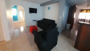 A modern living room with a black sofa, a red ottoman, and a flat-screen TV mounted on the wall.