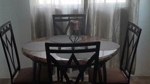 A round dining table with four chairs, a vase with a pink flower, and a window with sheer curtains.