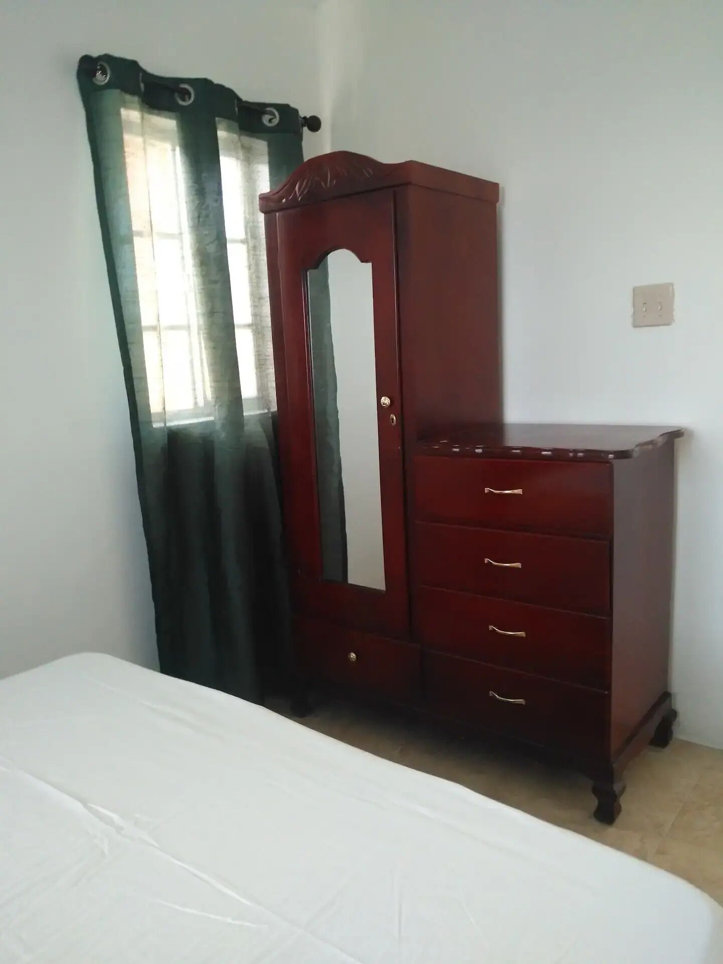 A room with a bed, a wooden dresser with drawers, a mirror, and a window with curtains.