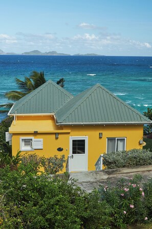 Ein gelbes Gebäude mit einem grünen Dach, einer weißen Tür und einem Fenster, in der Nähe von Wasser mit Blick auf das Meer und das entfernte Land.
