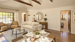Ein geräumiges Wohnzimmer mit hoher Decke, Holzbalken, einem großen Fenster, einem Sofa, einem Couchtisch und einem Essbereich, gedeckt mit weißem Geschirr.