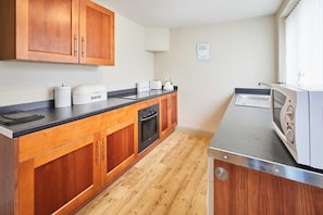 A kitchen with wooden cabinets, a black countertop, a built-in oven, a microwave, and a kettle. - Host & Stay | Moors Edge (Charltons)
