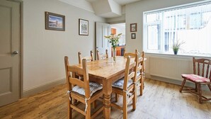 Une salle à manger avec une table et des chaises en bois, un vase rempli de fleurs et une fenêtre avec des stores.