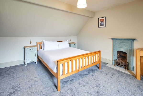 A bedroom with a wooden bed, white bedding, a nightstand, a dresser, and a fireplace.