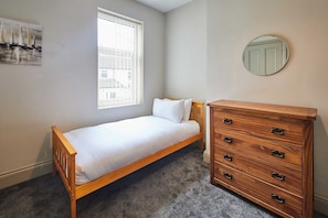 A small, neatly arranged bedroom with a single bed, a wooden dresser, a round mirror, and a window with blinds. - Host & Stay | Moors Edge (Charltons)