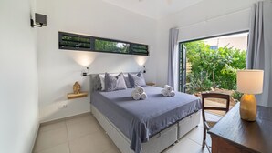 A bedroom with a bed, a ceiling fan, a window with curtains, a bedside table with a lamp, and a view of greenery outside. - Cute hideaway with private pool & garden. Close to nature & beaches. (Bonaire)