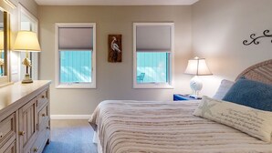 A bedroom with a bed, two windows with blinds, a dresser, and a lamp.