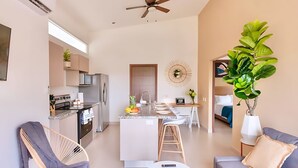 Un appartement moderne comprenant une cuisine, un coin repas et un salon. Il est équipé d’un ventilateur de plafond, d’une grande plante et d’un canapé.