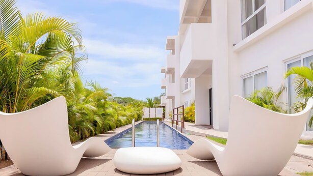 Un espace piscine doté de chaises blanches modernes, entouré de palmiers et bordé d’un bâtiment blanc avec des balcons.