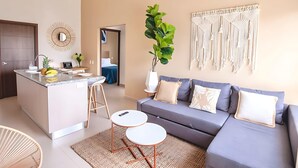 Un salon moderne avec un canapé gris, un petit îlot doté d’un évier et une décoration murale en macramé.