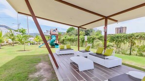 Un espace extérieur aménagé, abrité et doté de mobilier moderne, avec des canapés et des coussins blancs, une aire de jeux avec des toboggans, et une vue sur un quartier résidentiel verdoyant avec des bâtiments.