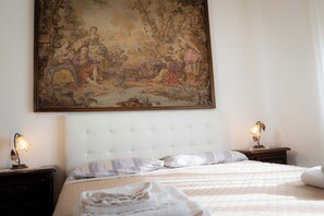 Un dormitorio con un cuadro clásico grande, un cabecero capitonado, dos mesitas de noche con lámparas y una cama bien hecha con almohadas.