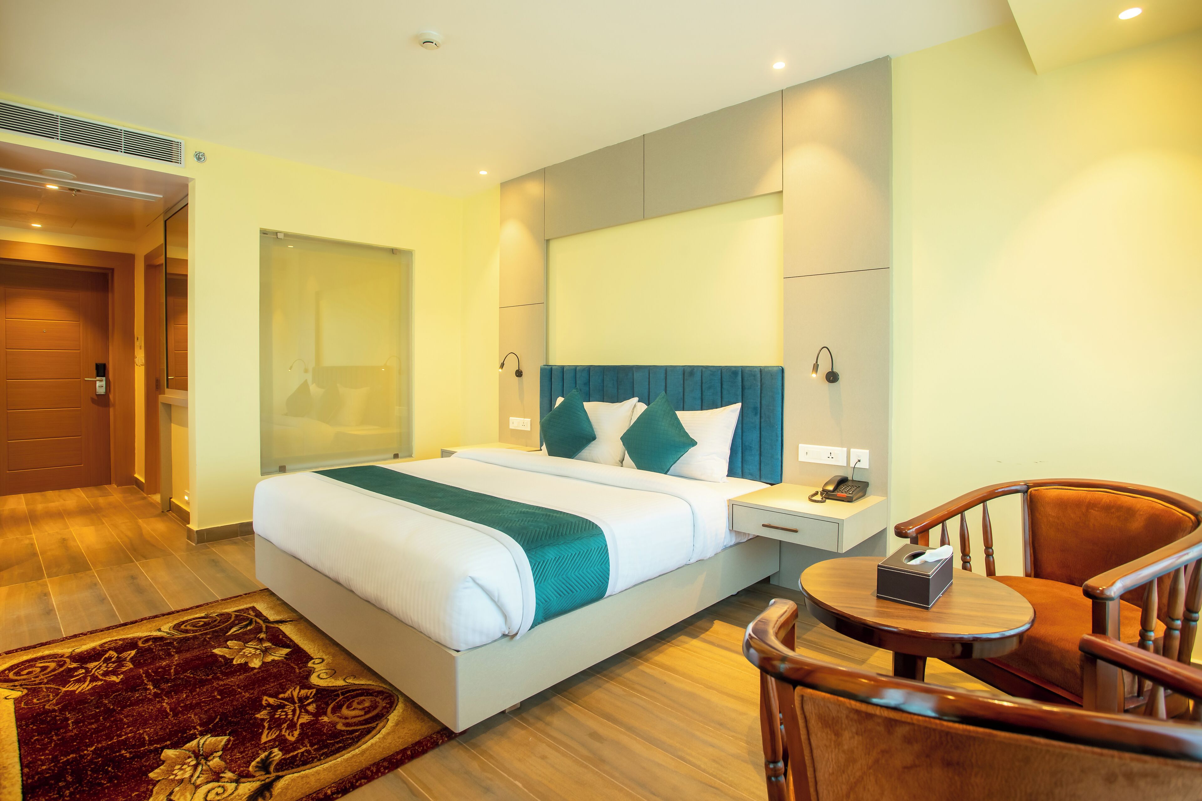 Habitación de hotel con una cama grande, mesitas de noche, una silla de madera y una pequeña mesa con una caja encima.