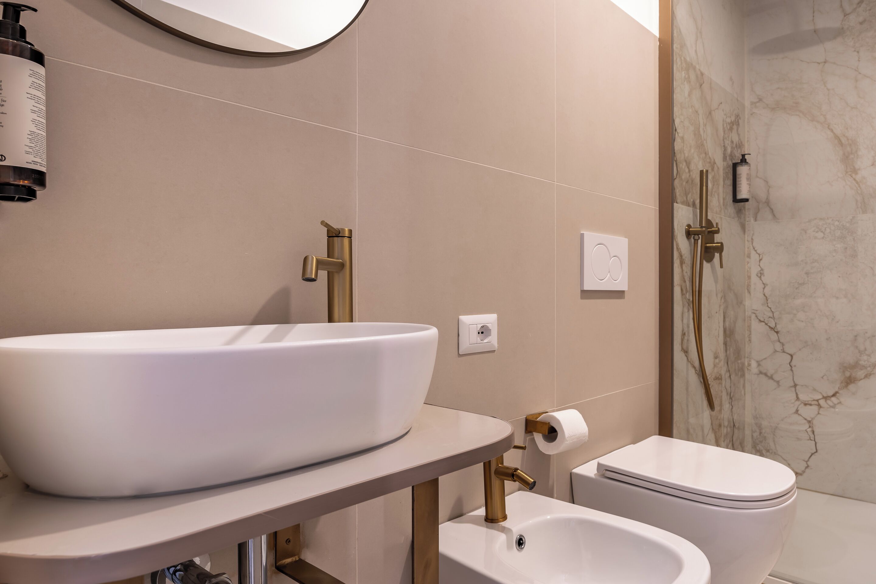 Ein modernes Badezimmer mit einem großen weißen Waschbecken, einem goldenen Wasserhahn, einer Toilette und einer Dusche mit Marmorwänden.