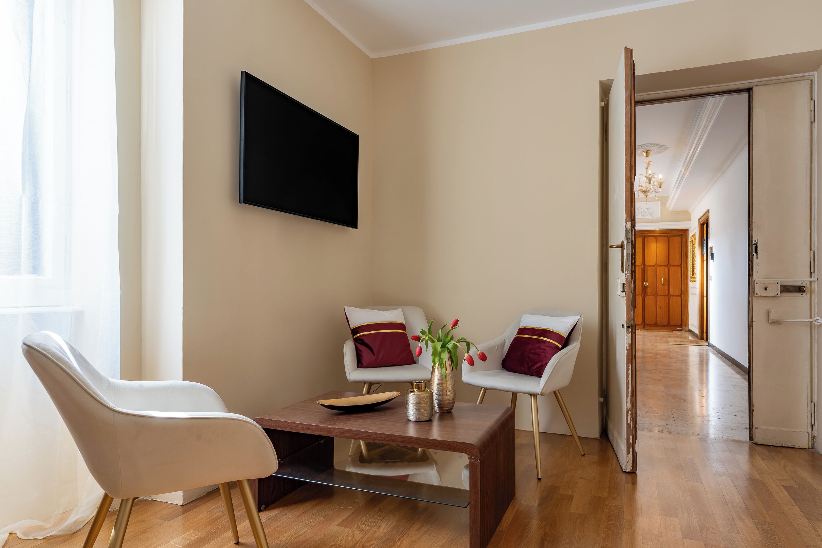 Una sala moderna con un televisor de pantalla plana, dos sillas blancas con cojines rojos, una mesa de centro y un jarrón con flores.