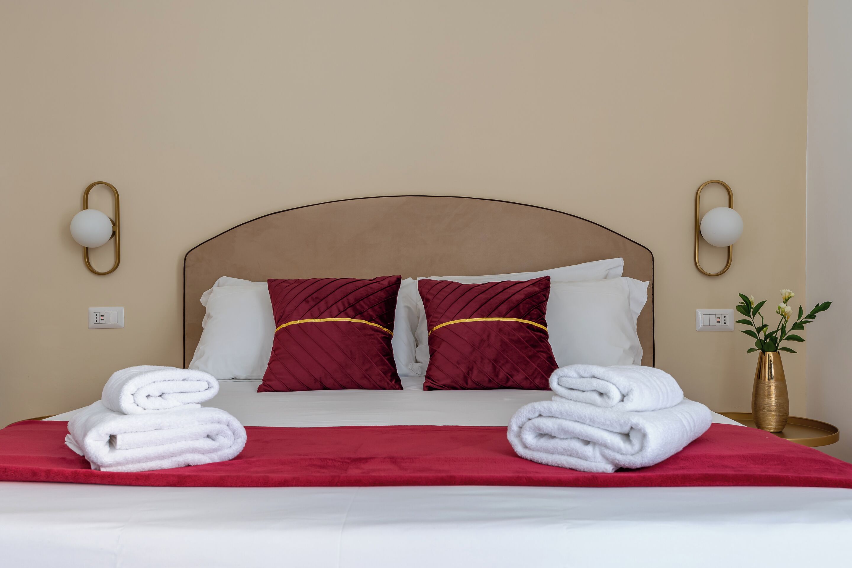 A neatly made bed with white and red bedding, two red pillows, and a gold vase with flowers on a bedside table.