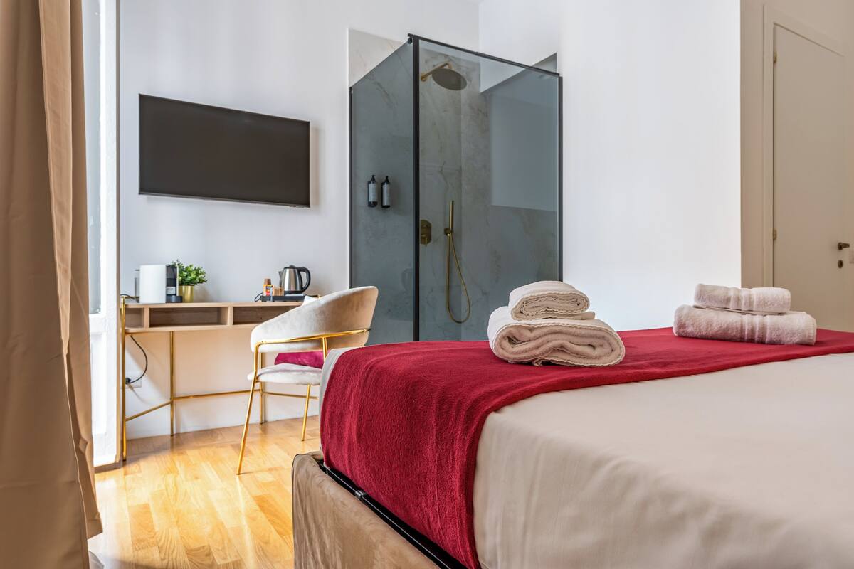 A modern hotel room with a bed, a desk, a chair, a shower area, and a mounted TV.