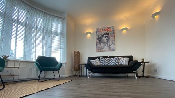 Een moderne woonkamer met een zwarte bank, een blauwgroene fauteuil en een groot raam met jaloezieën.