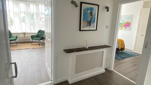 Een kamer met een houten vloer, een witte muur en een zwarte hanglamp. Er hangt een lijst met een afbeelding aan de muur en een klein plankje met een plantje.